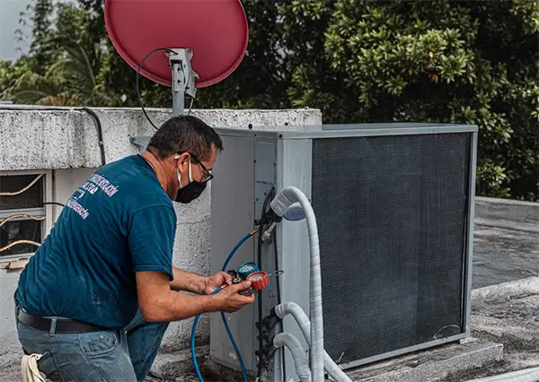 Technician repairing an air conditioner unit outdoors wearing a facemask and using a manifold gauge